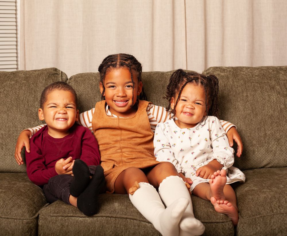 Three children laughing and playing together on a gray couch in casual clothing.