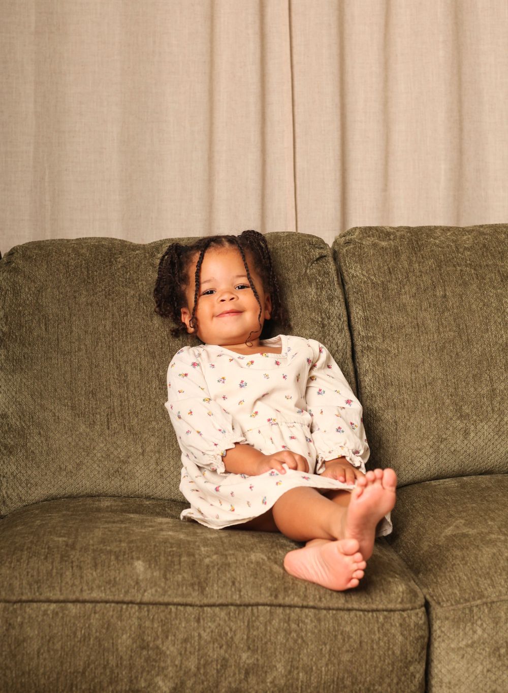 Young child in white patterned sleepwear sits comfortably on an olive green couch with a happy expression.