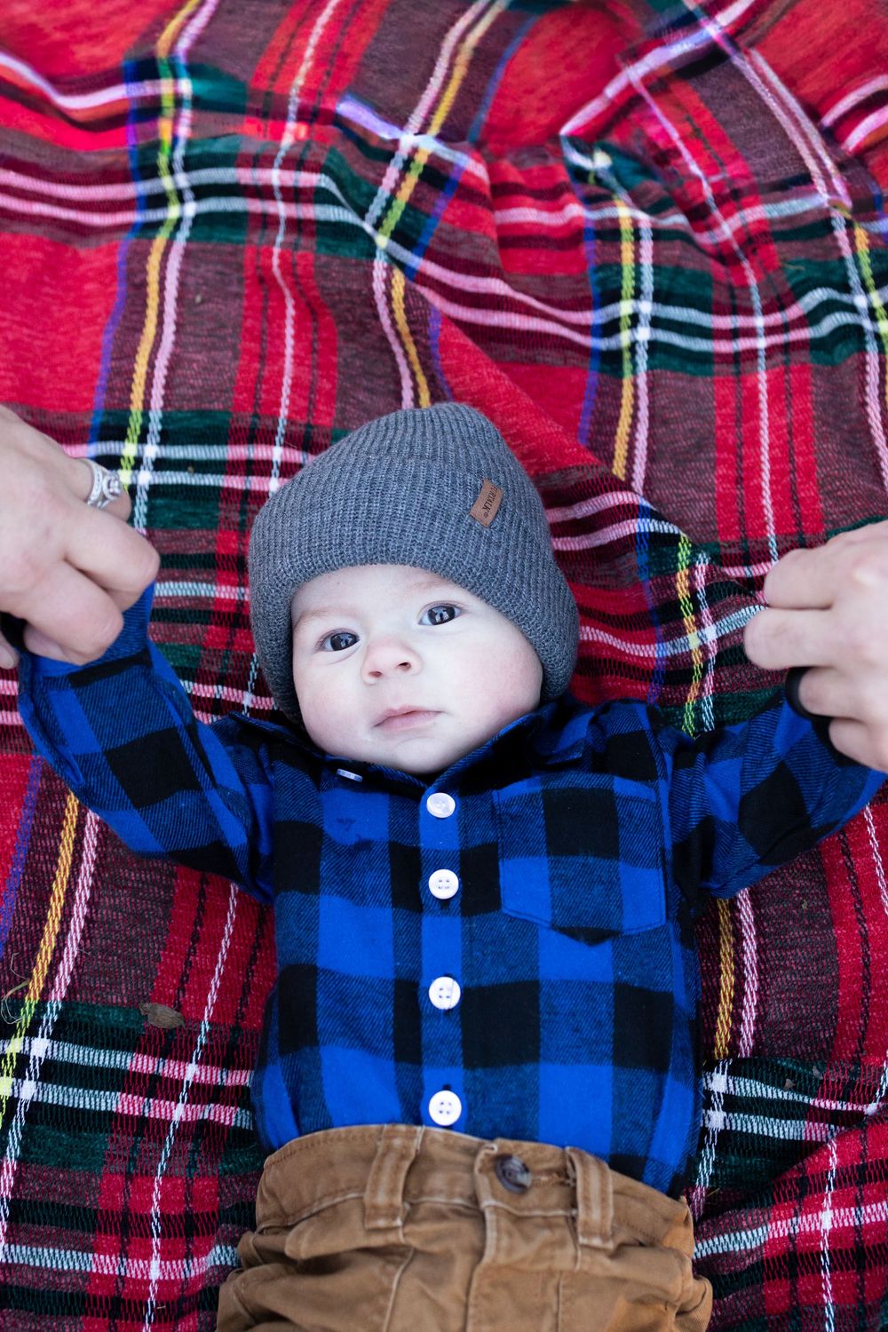 A baby wearing a gray beanie, blue plaid shirt and brown pants lies on a red plaid blanket.
