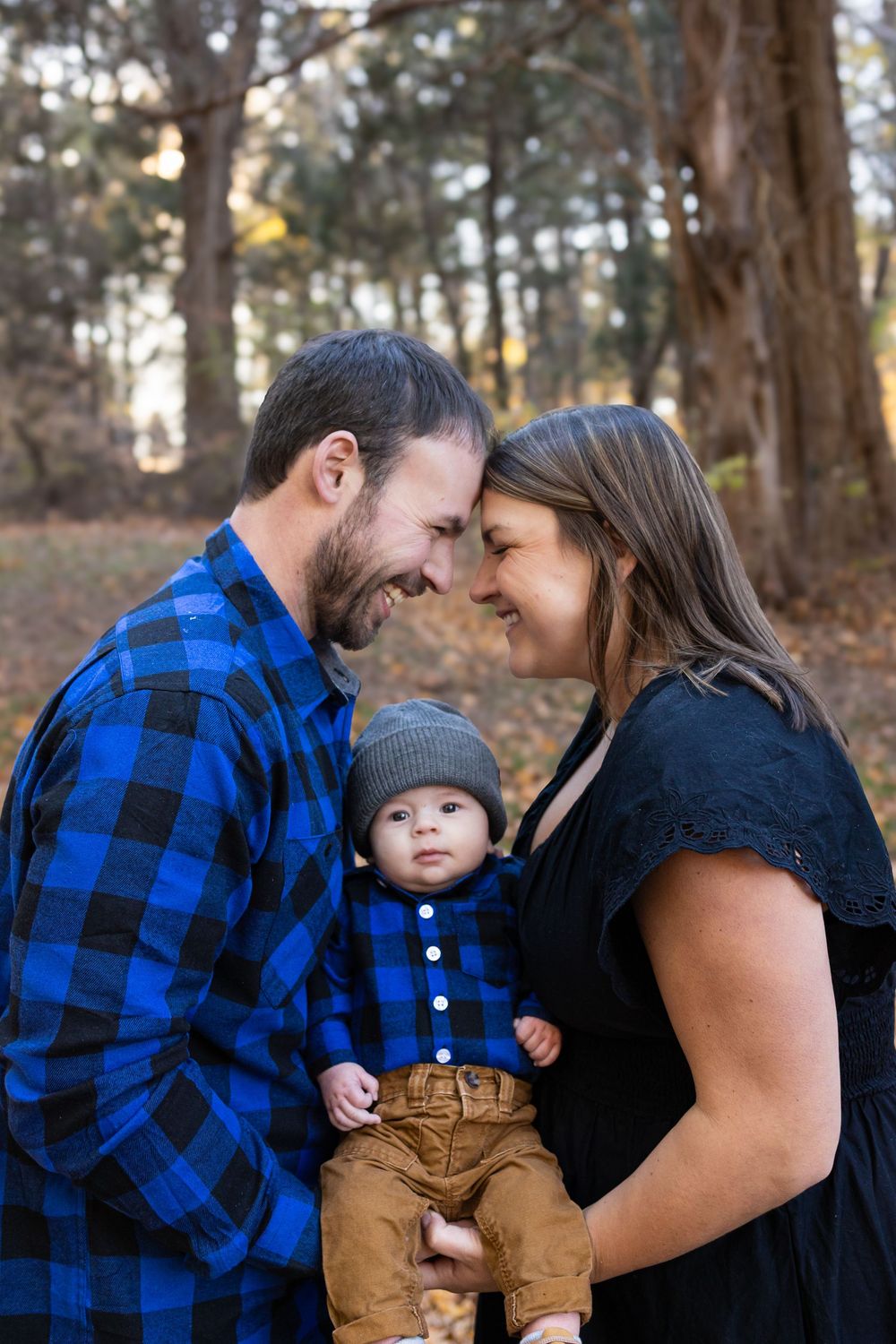 A couple shares a tender moment while holding their baby in brown overalls and beanie in a wooded outdoor setting.