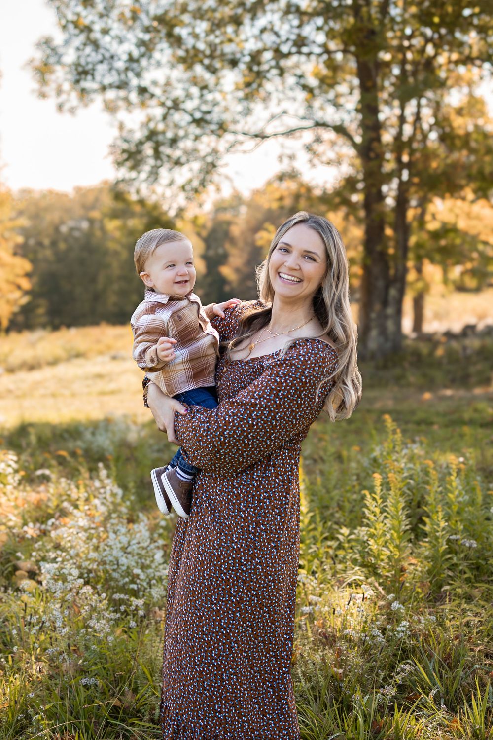 Mother and baby enjoying golden hour outdoors in a meadow with wildflowers and trees in the background. Fall Mini Session
