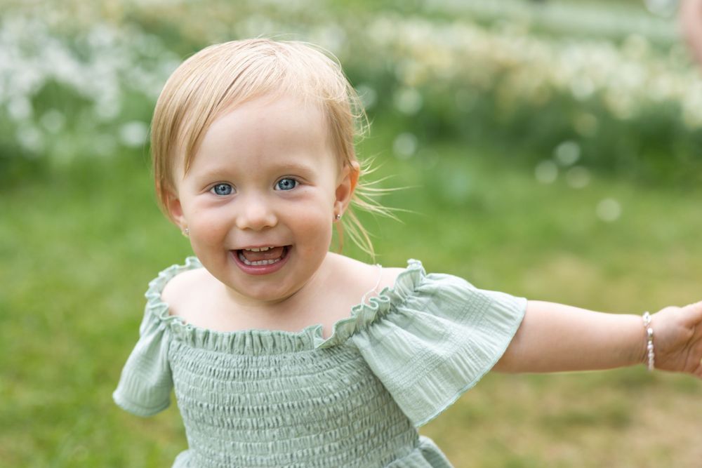 Toddler wearing a mint green smocked dress smiles joyfully while playing outdoors in a sunny garden setting. Spring Mini Session