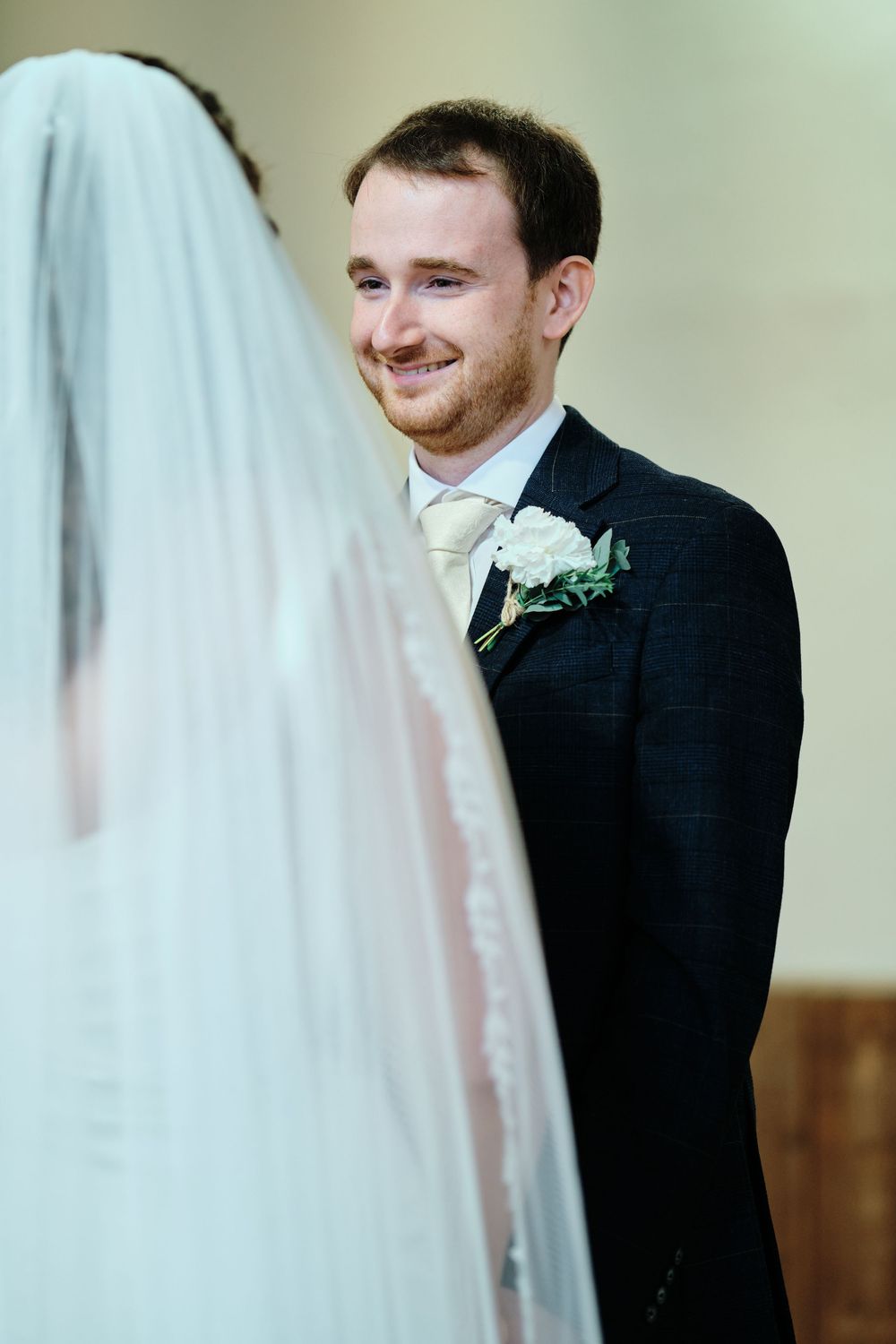 A groom in a dark suit with a white boutonniere smiles lovingly at his bride during their wedding ceremony.