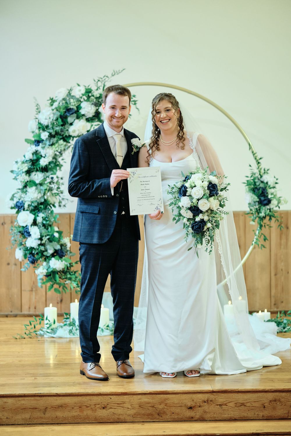 Newlyweds pose with their marriage certificate in front of a wooden wall decorated with white floral arrangements.