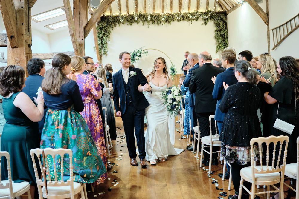 Wedding guests line the aisle of a bright indoor venue during a ceremony with white chairs and floral decorations.