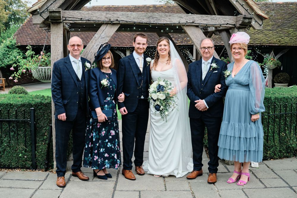 A formal wedding party poses together outside a rustic stone church entrance on a sunny day.