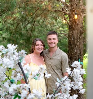 A couple poses together outdoors near a tree with white blossoms in a romantic garden setting.