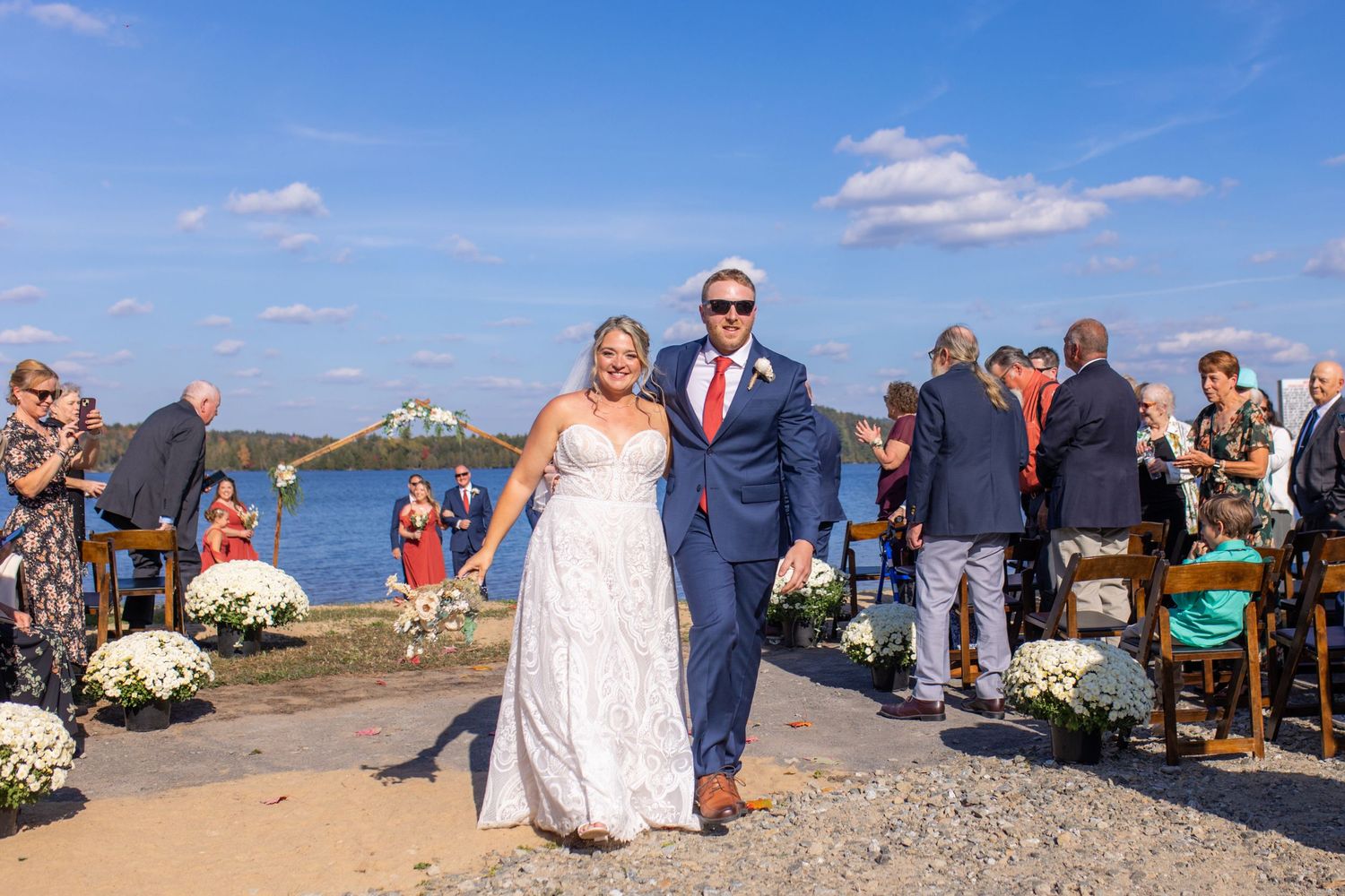 Outdoor beach wedding ceremony with guests gathered on the rocky shoreline under blue skies.