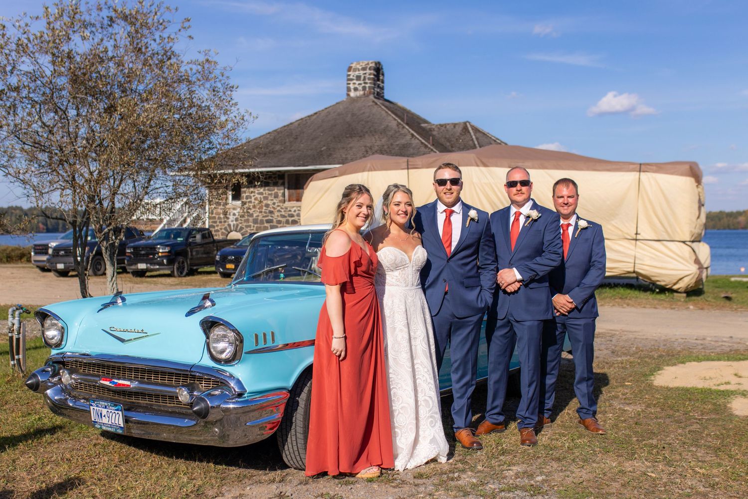 Wedding party poses with vintage blue car in front of rustic building wearing navy and coral attire.