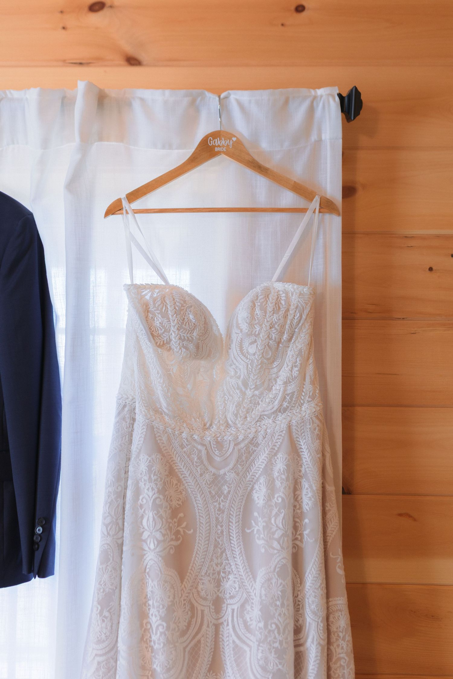 A white lace wedding dress hangs on display against a rustic wooden wall.