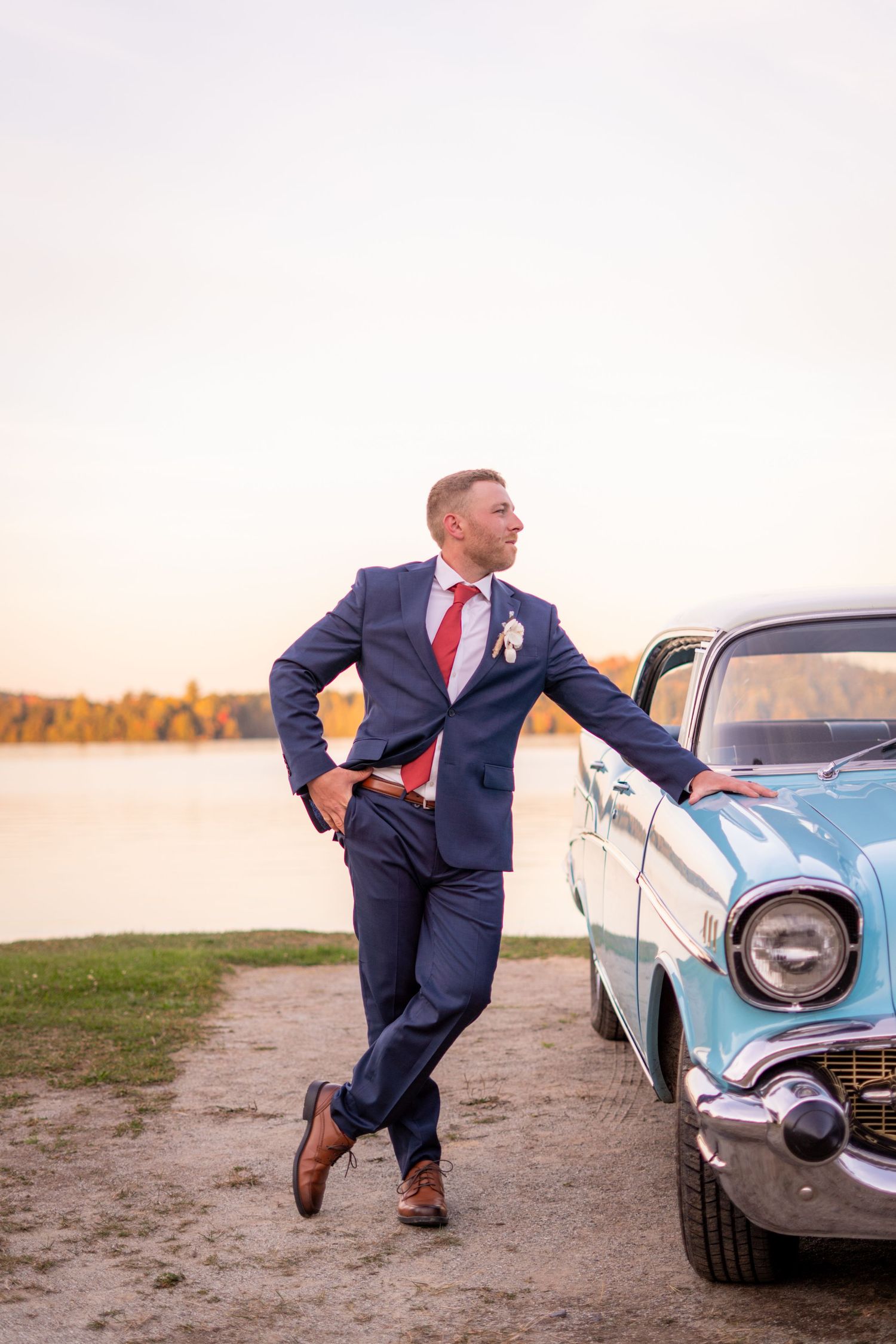 Groom in navy suit poses playfully with vintage blue car.