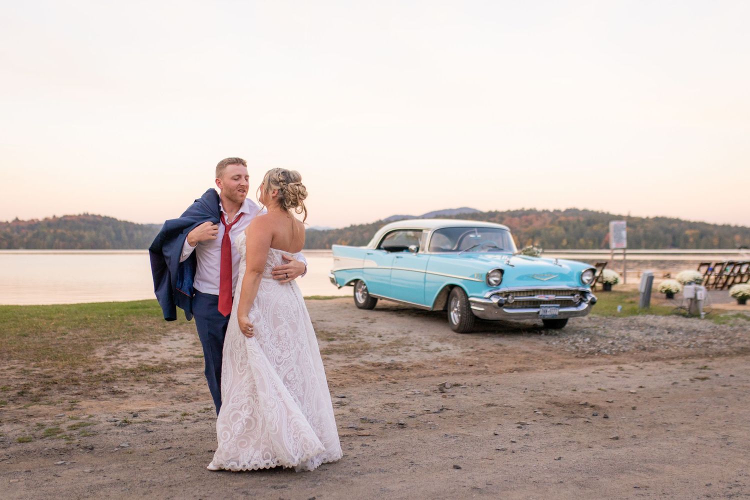 Newlyweds walking near vintage blue car at sunset by waterfront.