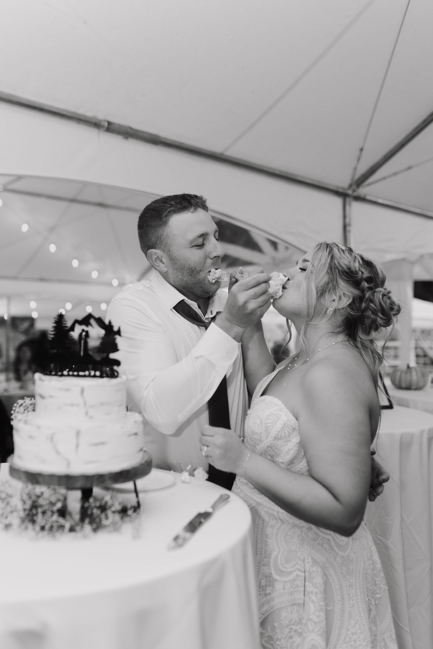 A couple in wedding attire share a playful moment while cutting their white wedding cake at a tented reception.