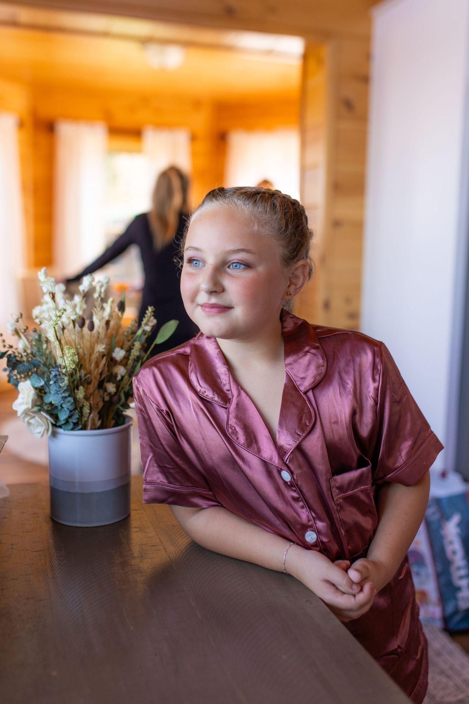 A child in a mauve satin robe sits at a table with a floral arrangement in soft morning light.