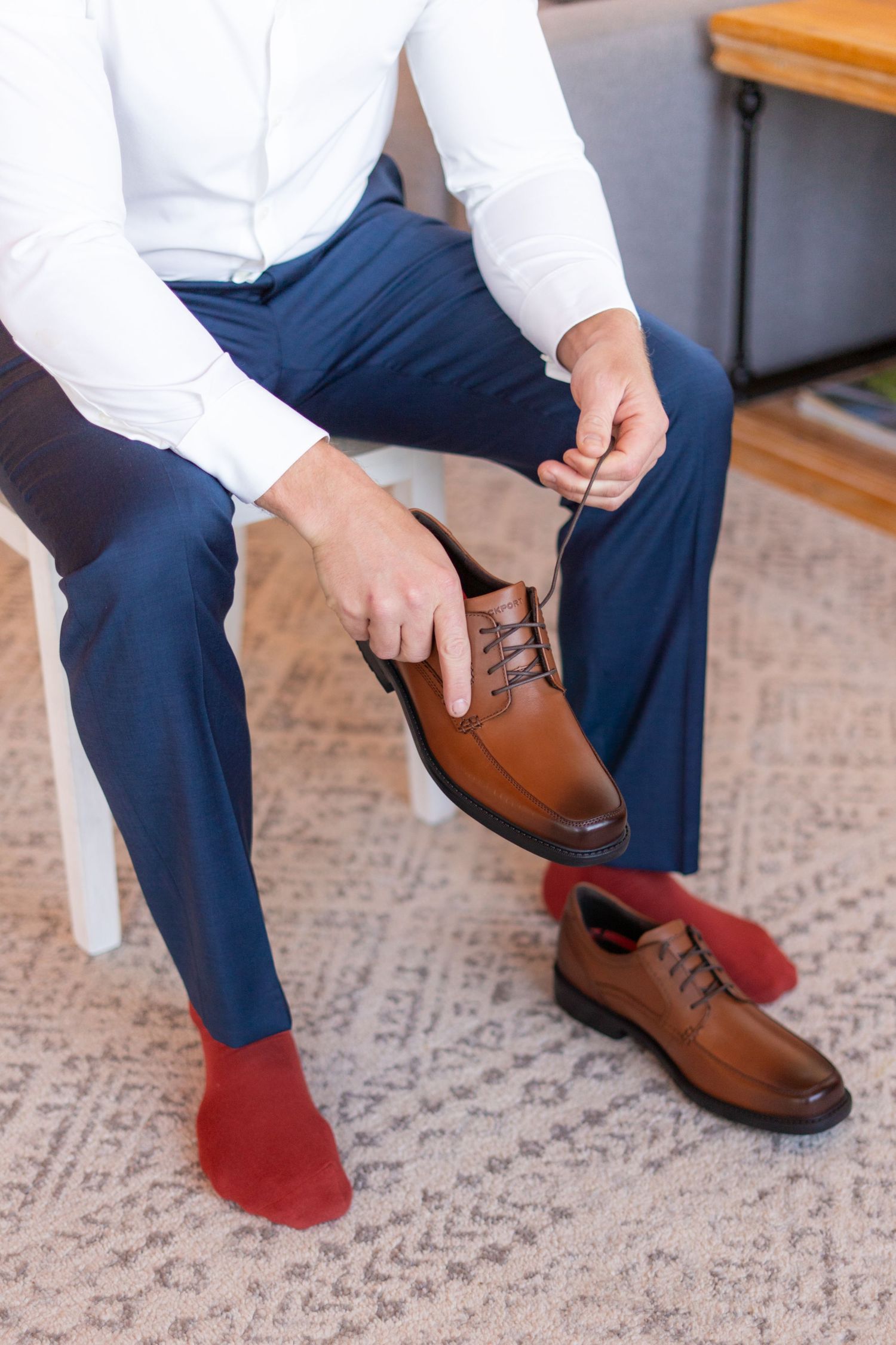 Man in navy pants and white shirt putting on brown leather dress shoes with red socks while sitting.