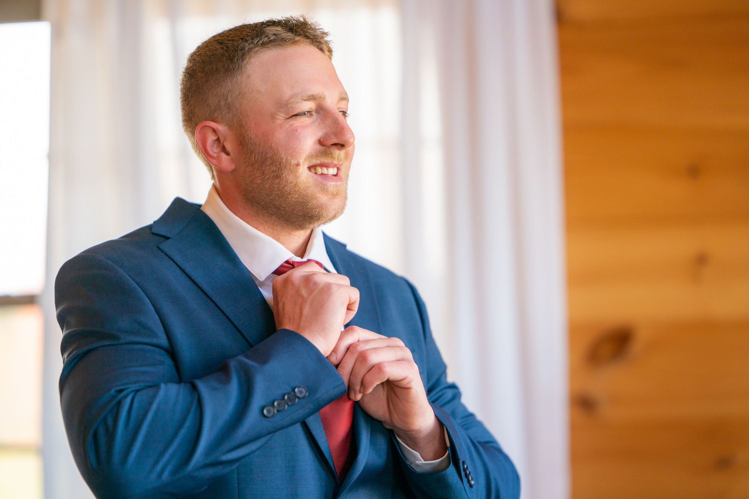 A person wearing a navy blue suit and red tie smiles while seated in front of a wooden wall.