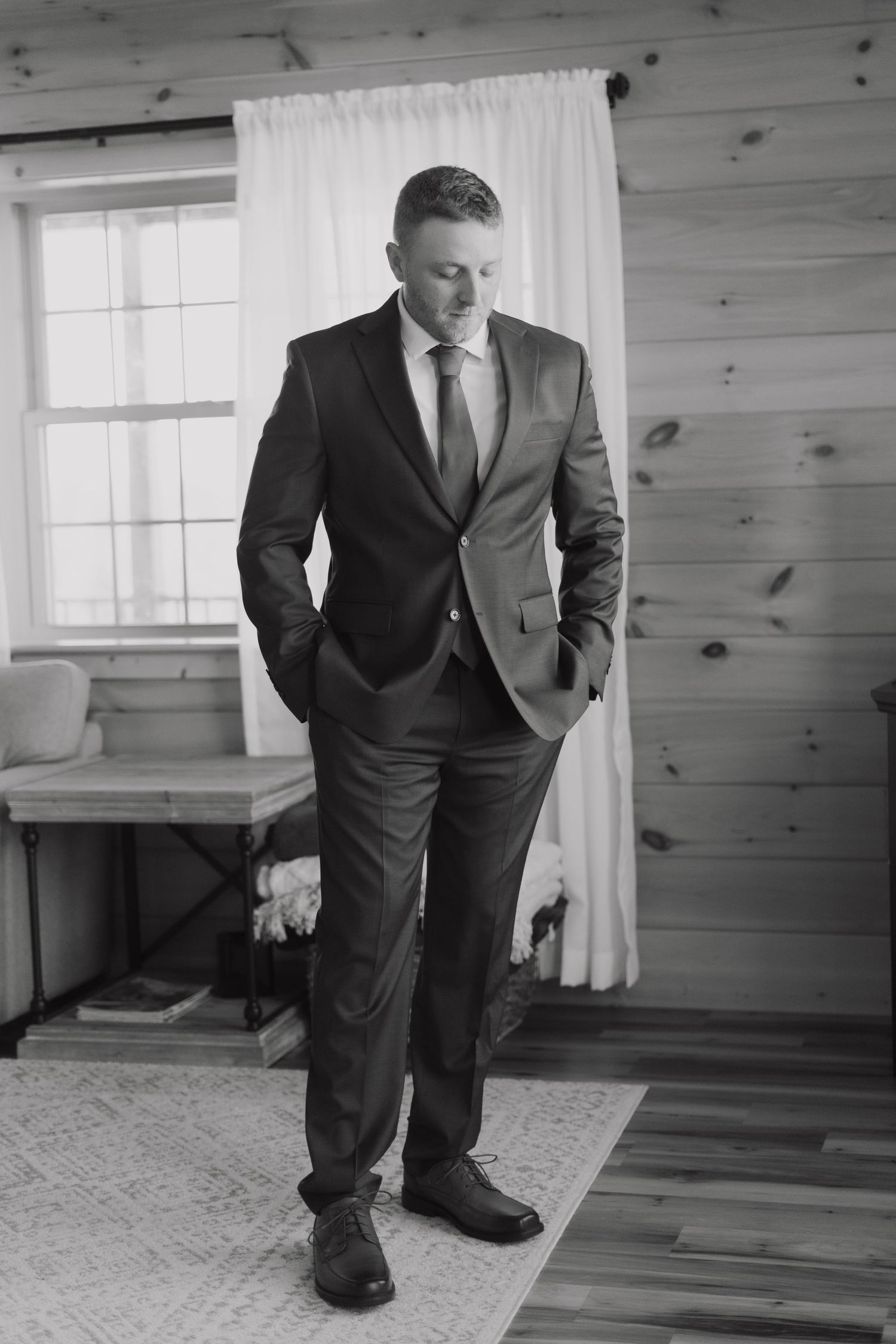 Black and white portrait of a groom in a formal suit standing by a window in a rustic wooden room.