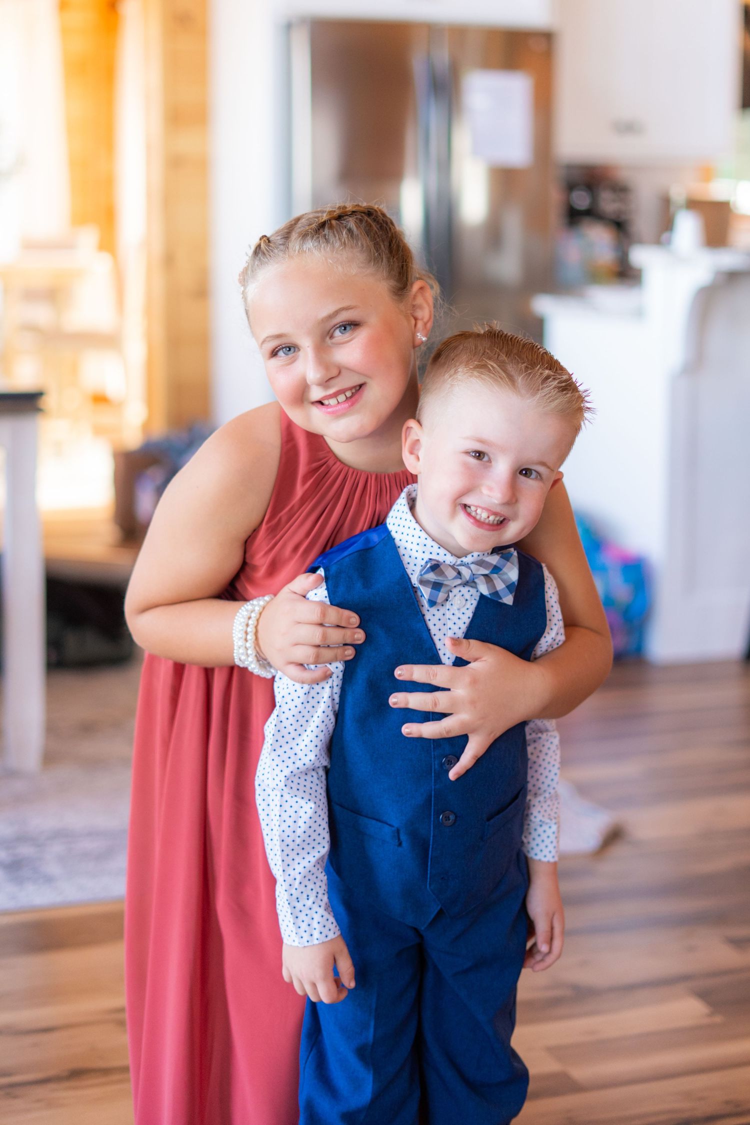 Young child in a navy blue vest and bow tie being hugged in a sunlit room.