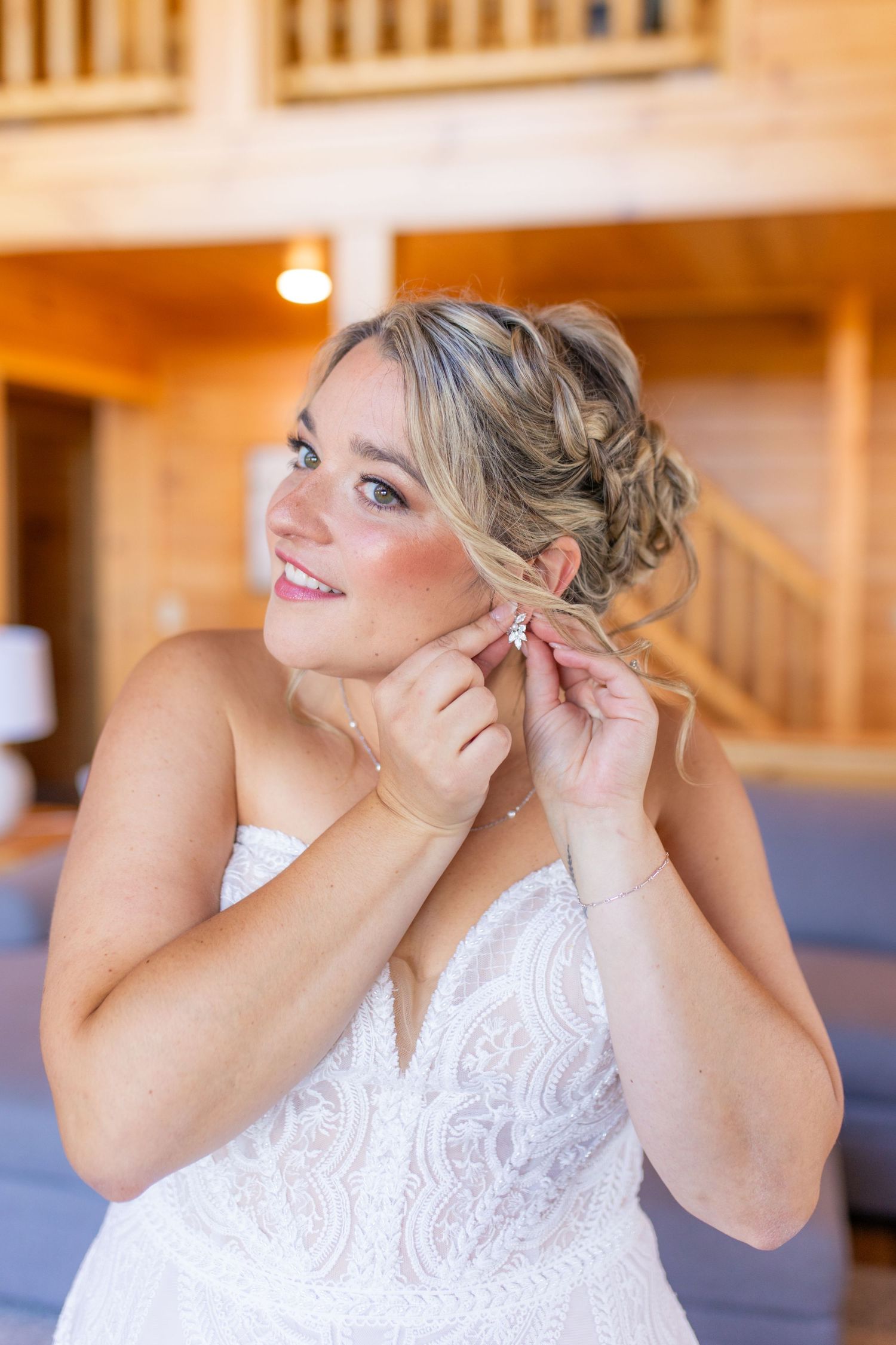 Bride in white lace dress putting on earrings while getting ready in a cabin setting.