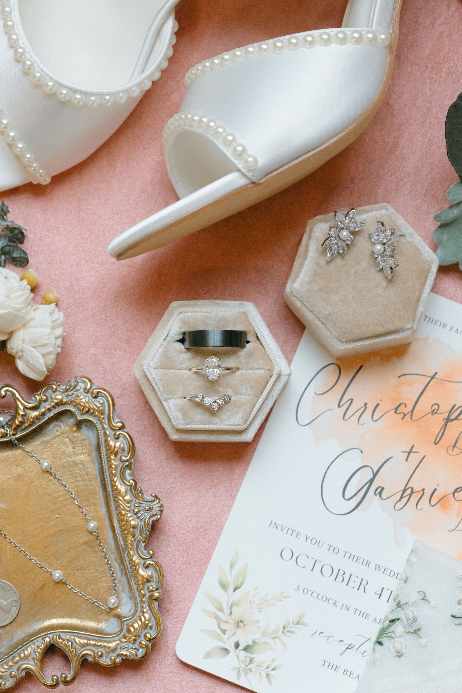 Elegant wedding details including white satin shoes, pearl jewelry, and calligraphy invitations on a blush pink background.