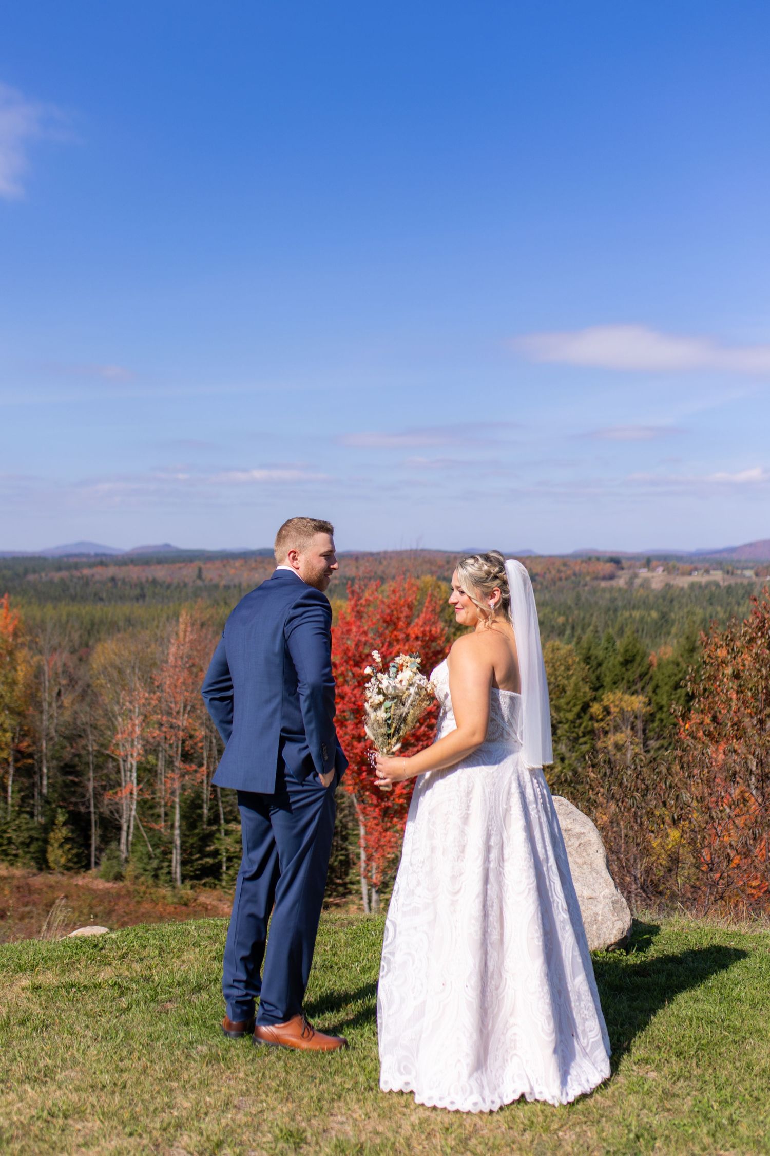 Couple in wedding attire standing on hilltop overlooking autumn mountain landscape.