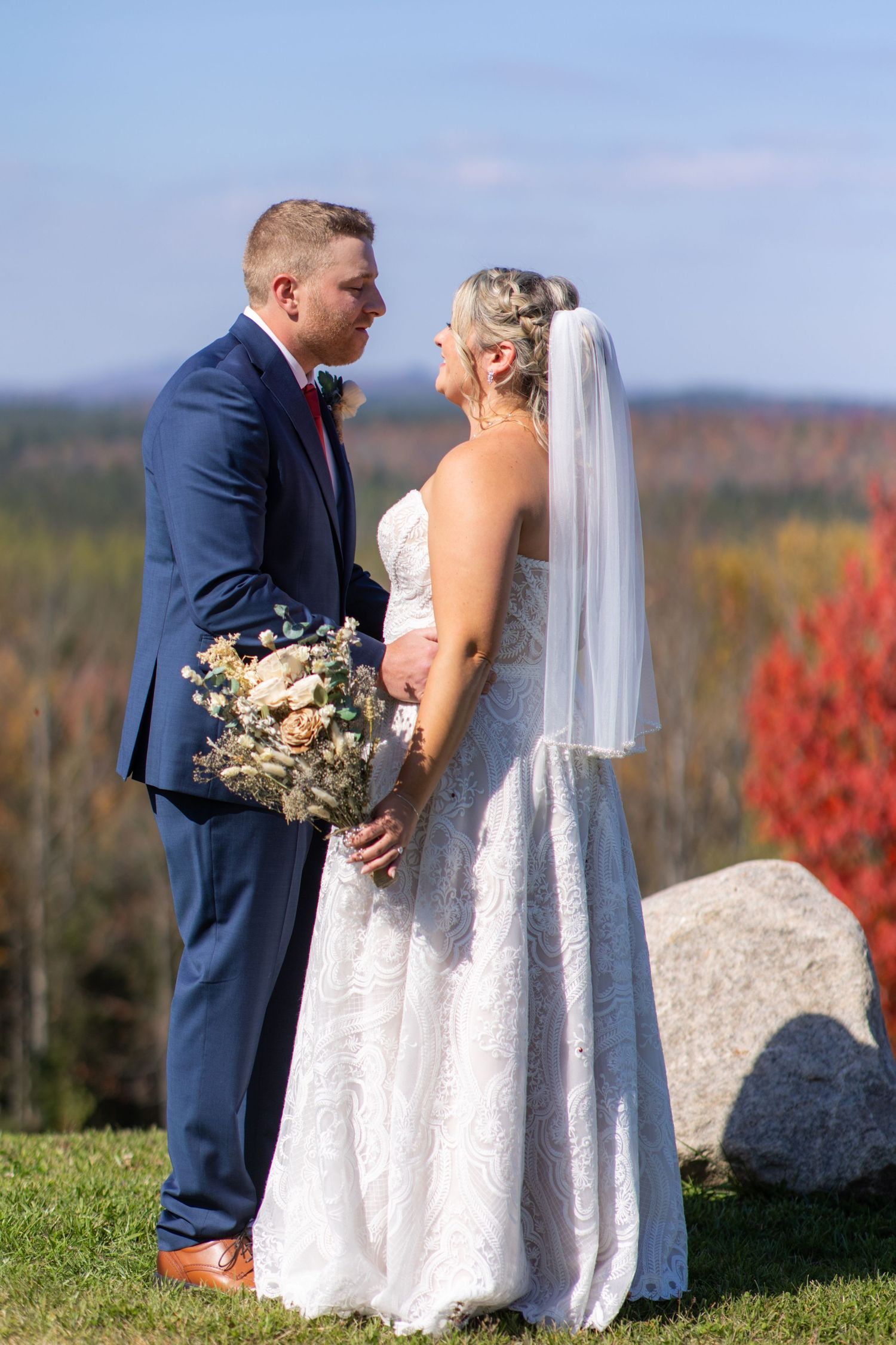 Bride and groom share intimate moment against autumn landscape with red foliage.