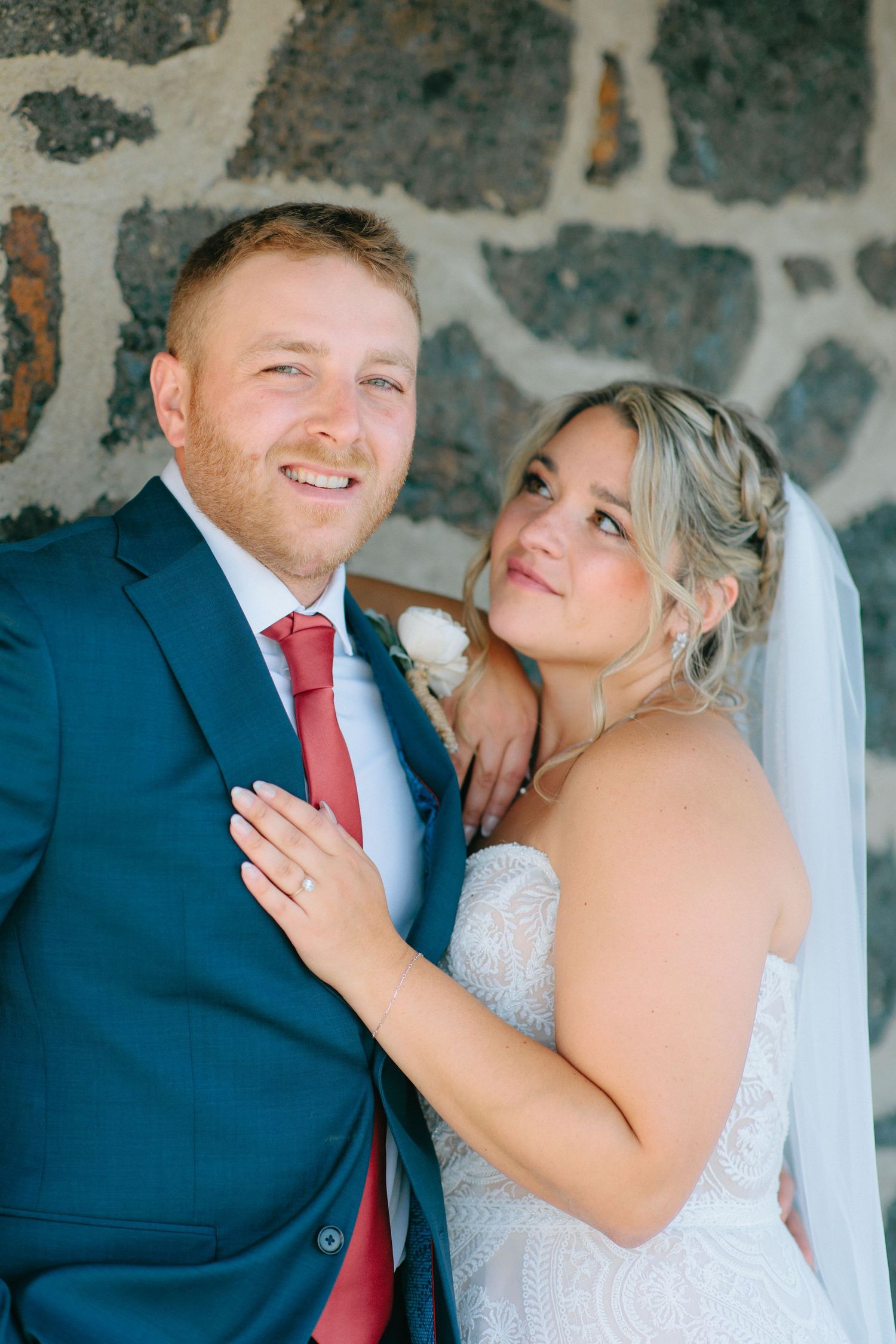 Wedding couple in navy suit and white dress posing against a stone wall.