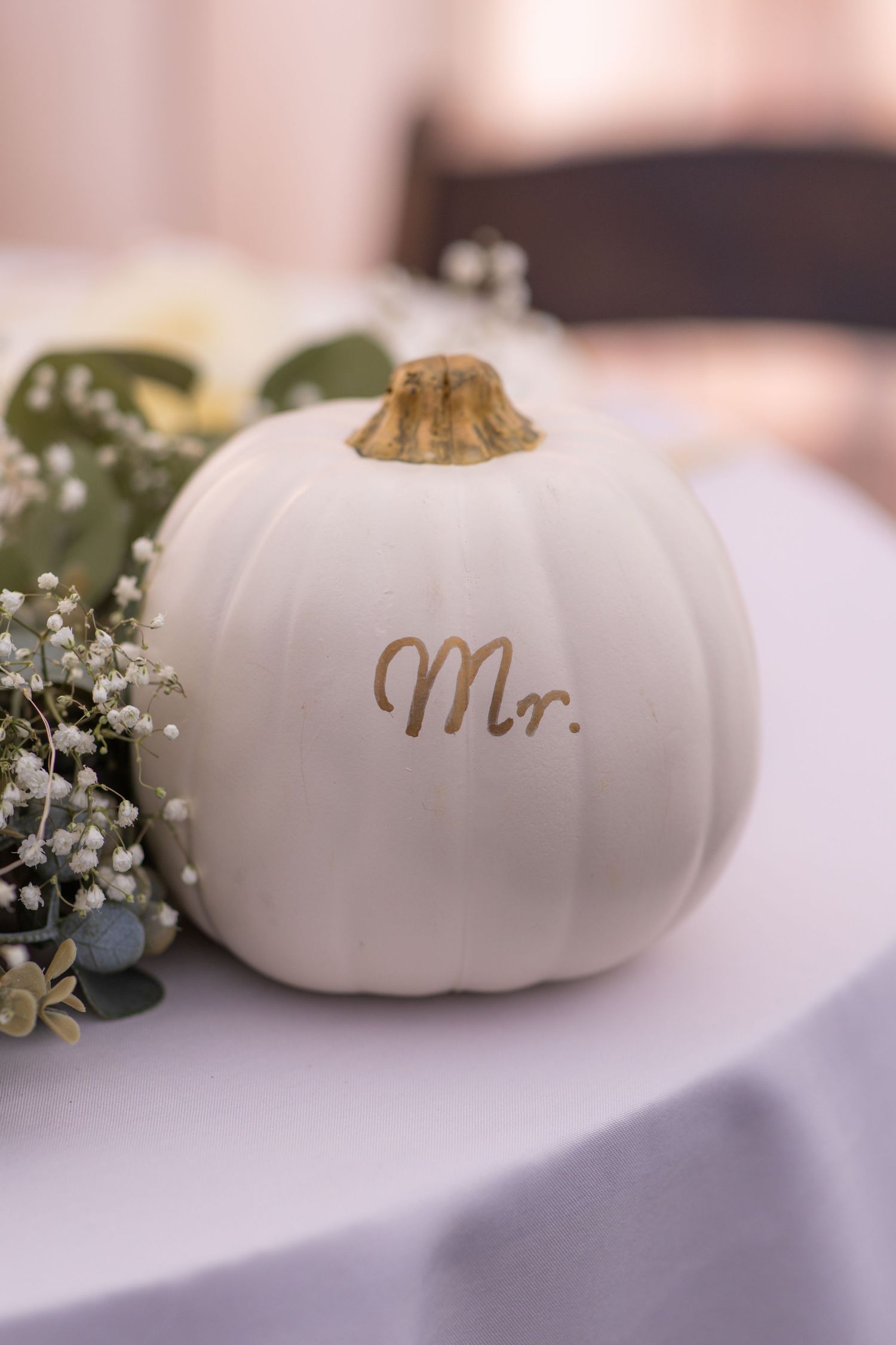 White decorative pumpkins with gold lettering arranged with baby's breath flowers on a table.