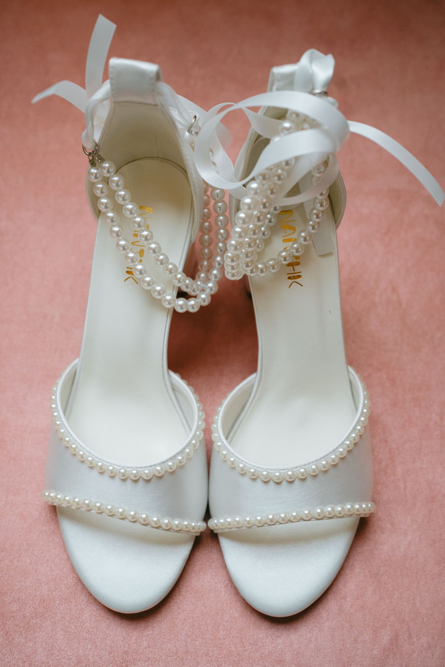 Elegant white bridal shoes adorned with delicate pearl details displayed on a pink background.