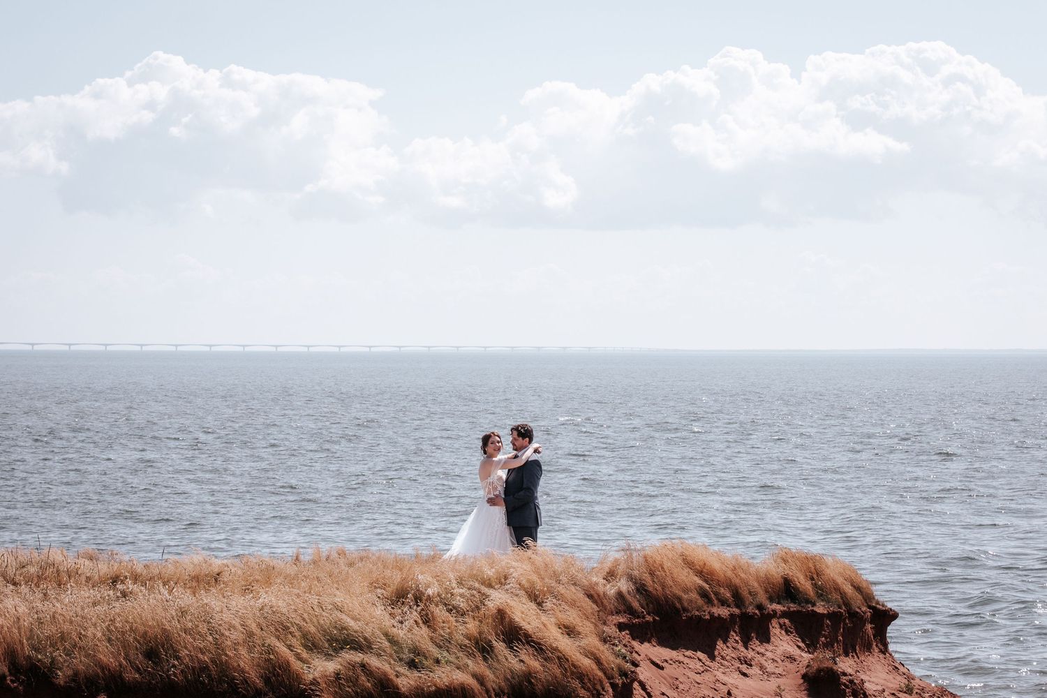 Two people embrace in silhouette against a scenic coastal landscape with tall grass and ocean views at sunset.