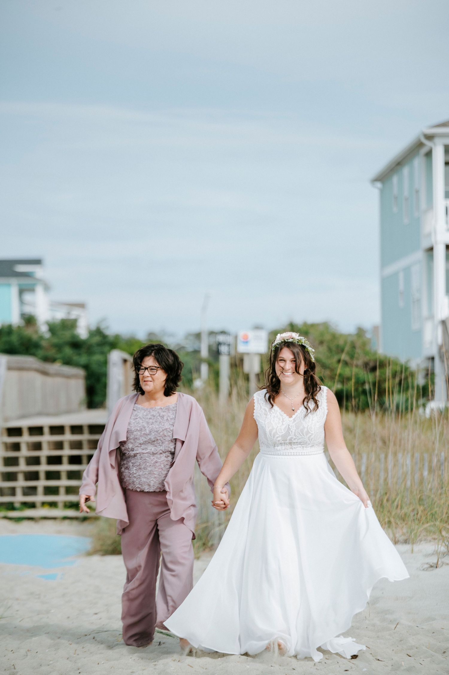 Two people in wedding attire walk together on a sandy beach with coastal buildings in the background.