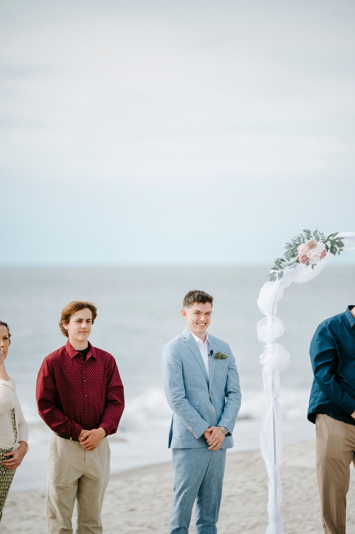 Groomsmen lined up on a beach striking playful poses in burgundy sweaters and gray suits against an overcast sky.