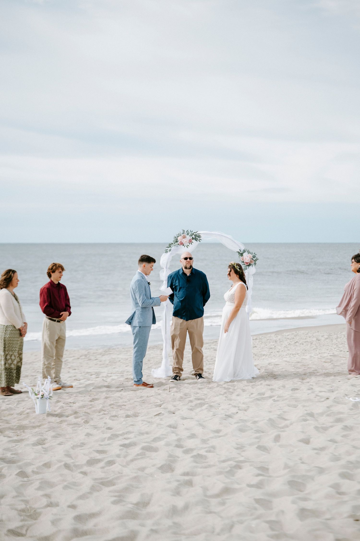 Wedding guests in pastel outfits stand in a line on a beach during a seaside ceremony.