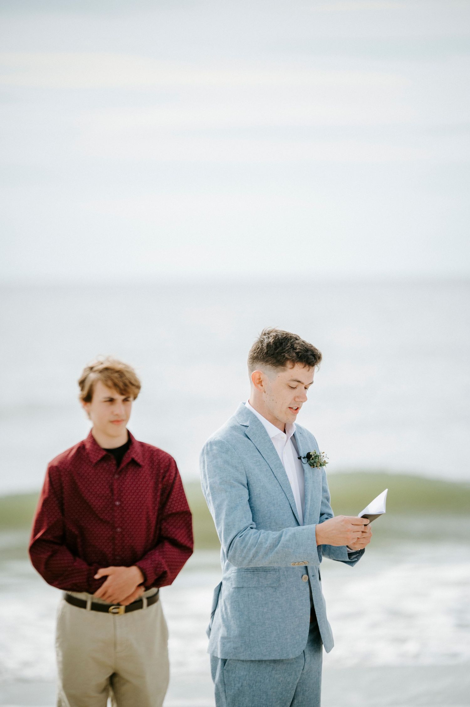 Two people stand on a beach wearing burgundy and light blue outfits while reading papers by the ocean waves.