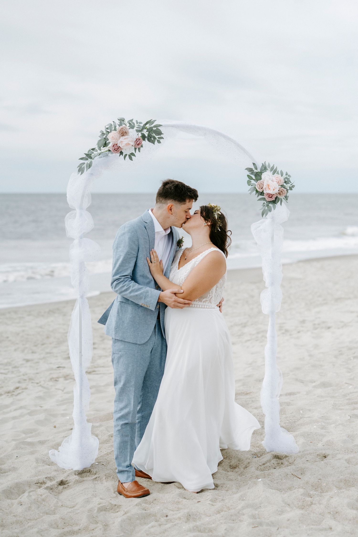 A romantic beach wedding ceremony with a white floral arch and couple embracing on the sand by the ocean.