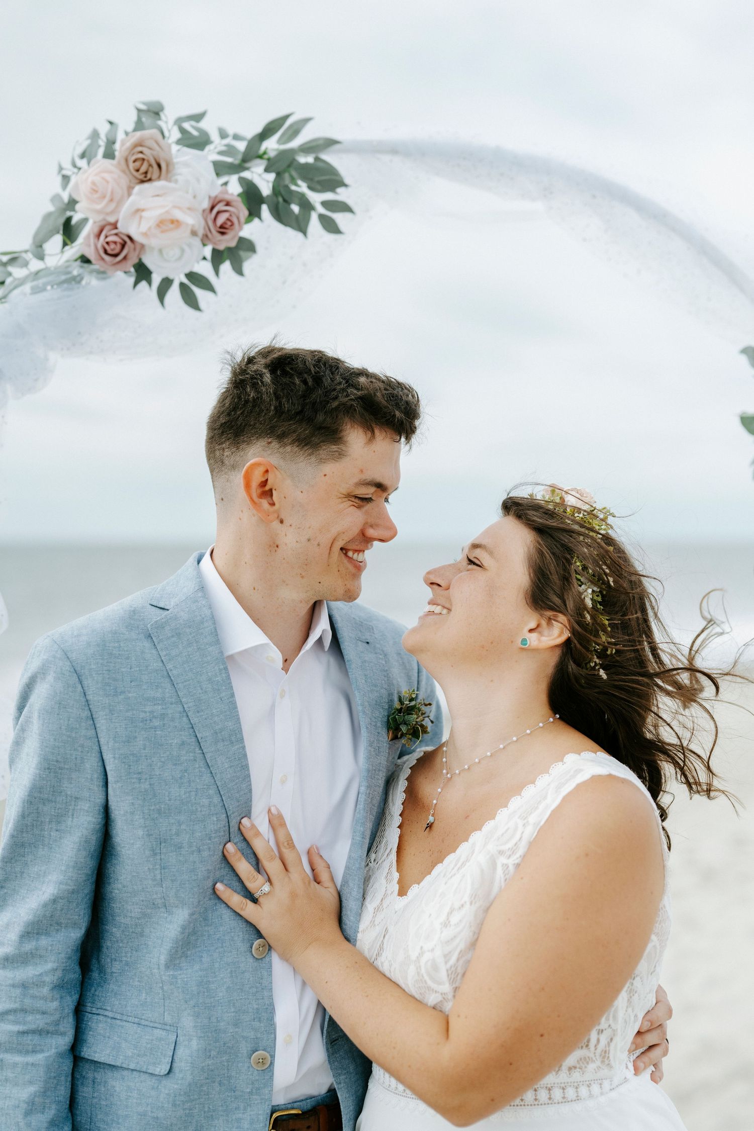 A romantic wedding couple shares intimate moments on a beach with soft floral decorations and overcast skies.