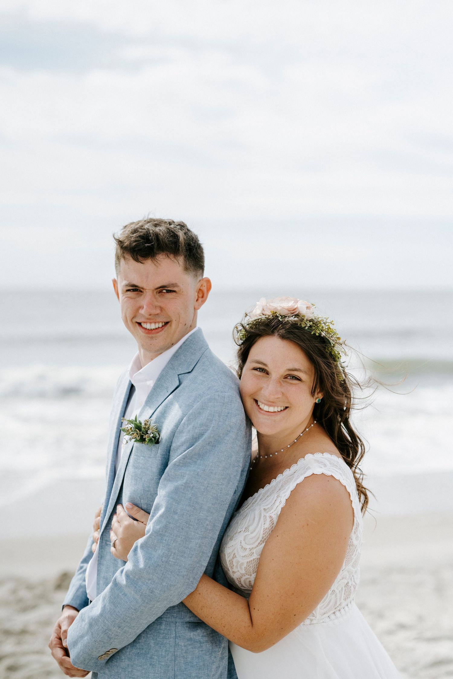 A joyful wedding couple poses together on a beach with waves and cloudy skies in the background.