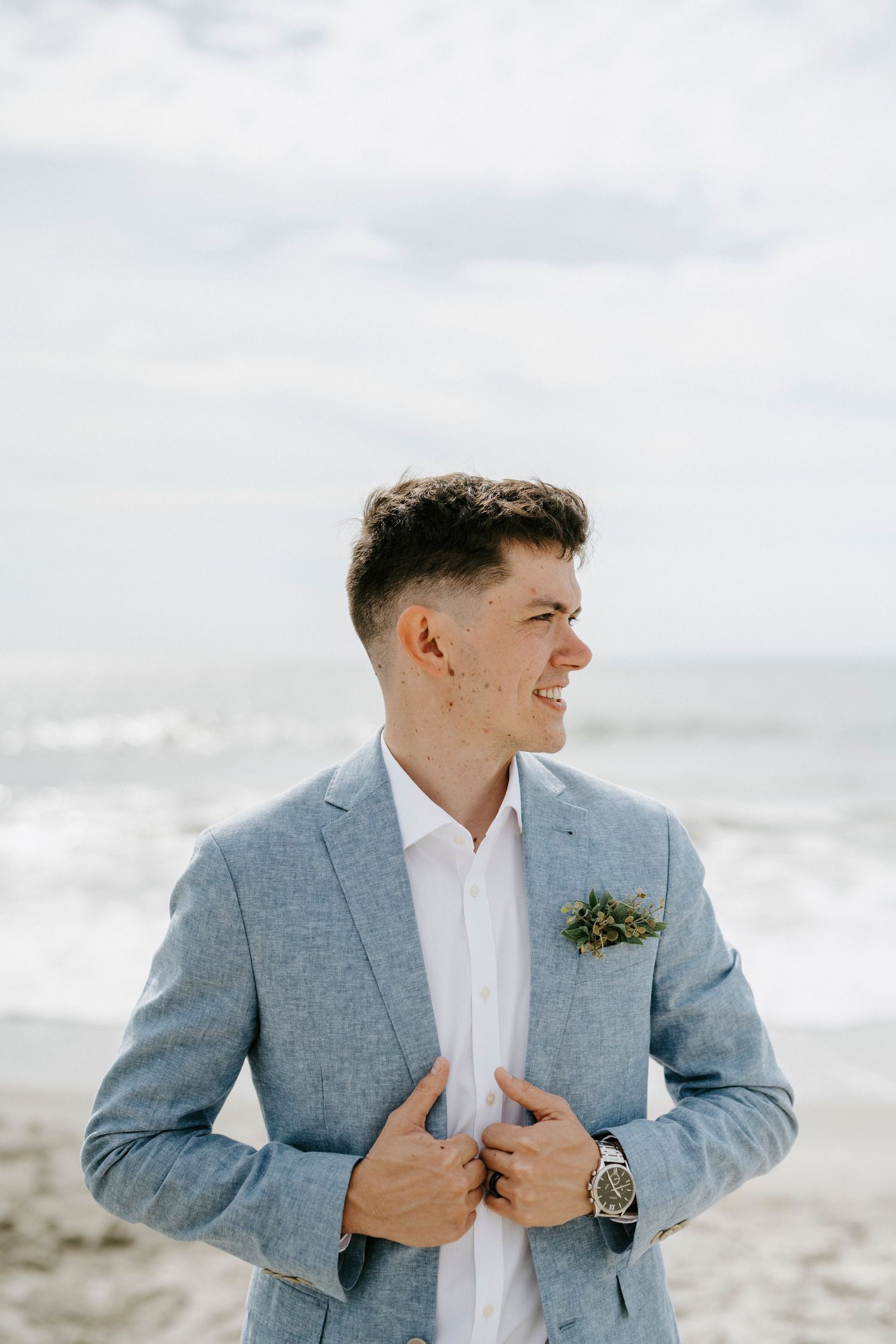 Groom in light gray suit and white shirt poses against cloudy beach backdrop.
