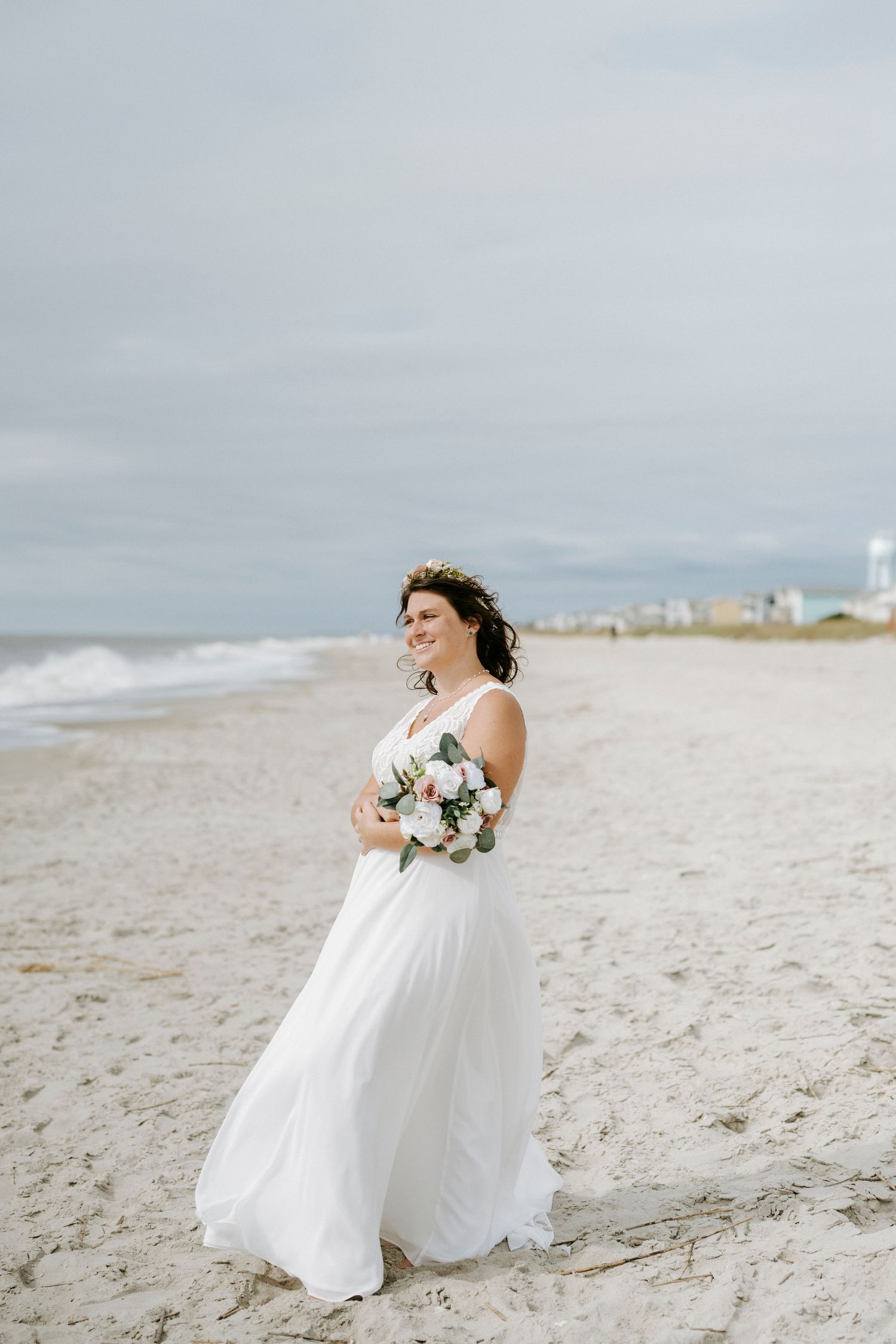Bride in flowing white dress poses with bouquet on serene beach landscape.