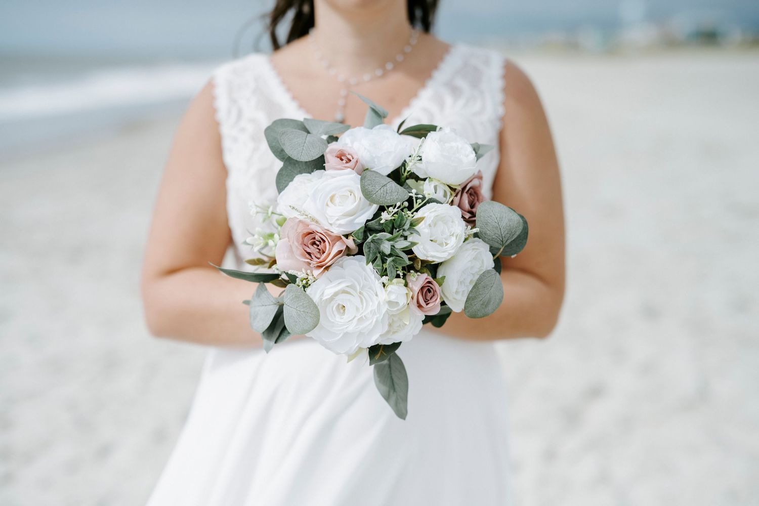 A delicate bridal bouquet with white and blush pink roses mixed with eucalyptus leaves held against a white dress.