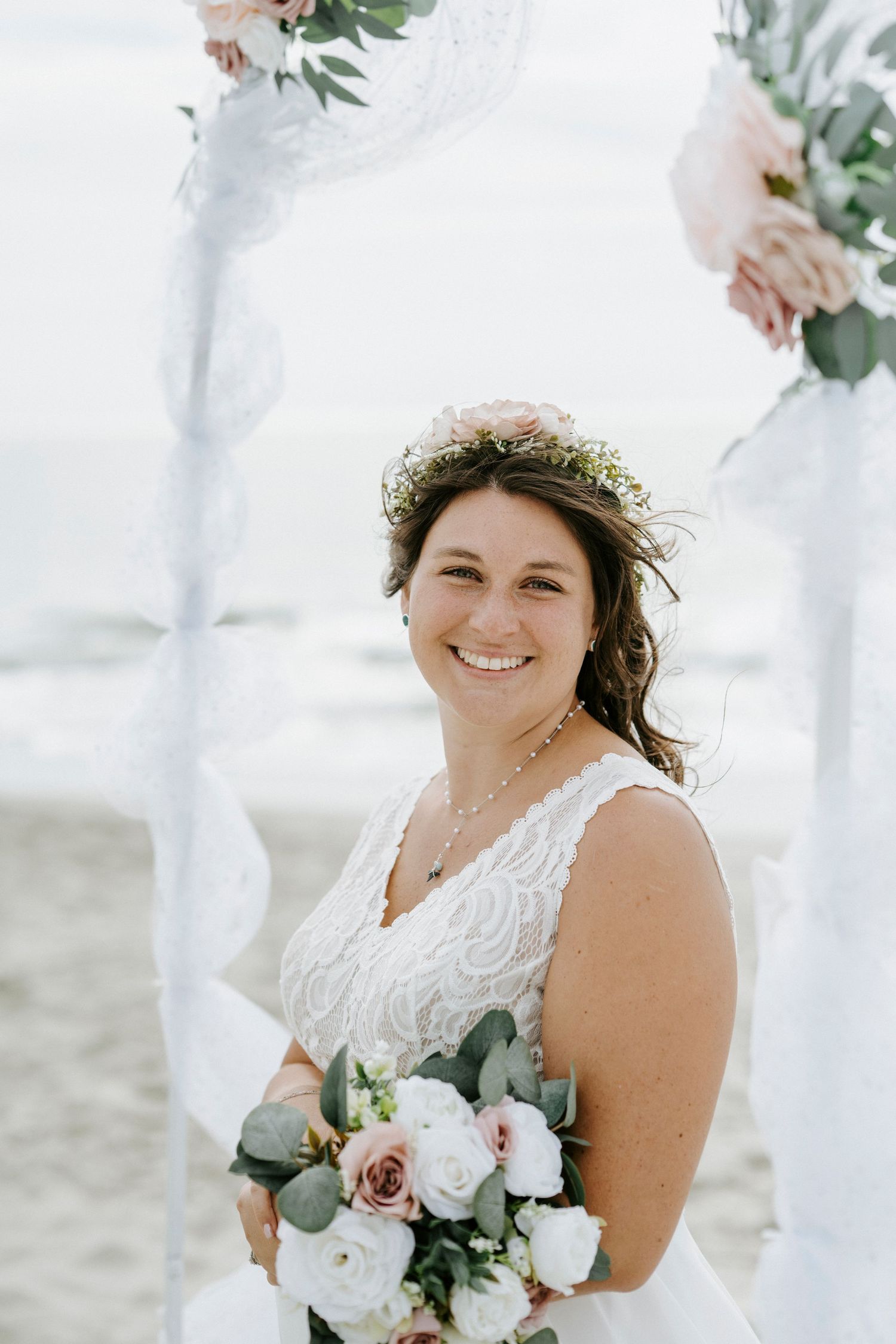A bride in a white lace dress wears a floral crown and holds a bouquet with roses and eucalyptus.