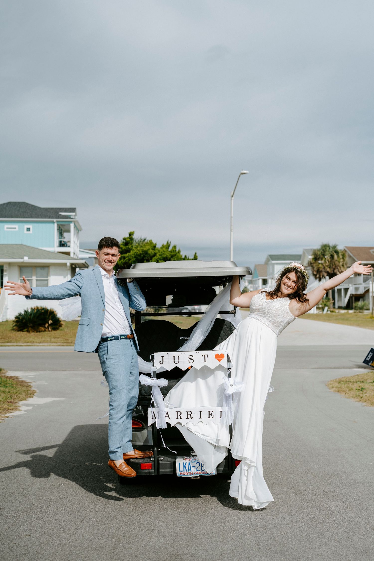 Newlyweds pose playfully with their decorated getaway car in a coastal parking lot.