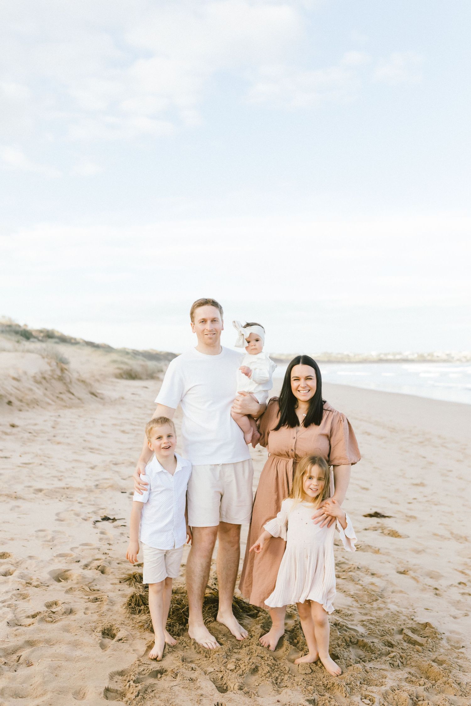 A family group sits together on a sandy beach wearing coordinated neutral-toned outfits during a sunset photoshoot.