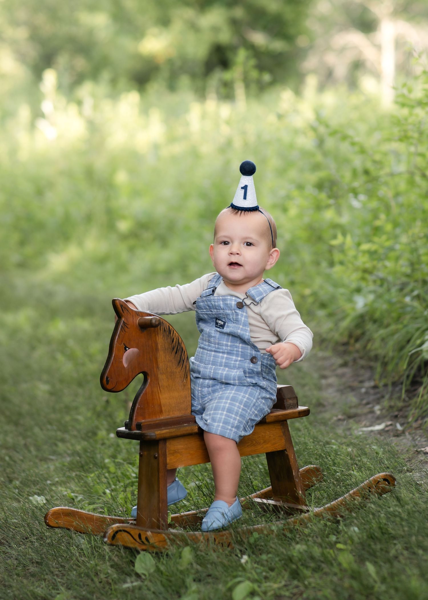A baby wearing blue overalls sits on a wooden rocking horse in a grassy outdoor setting.