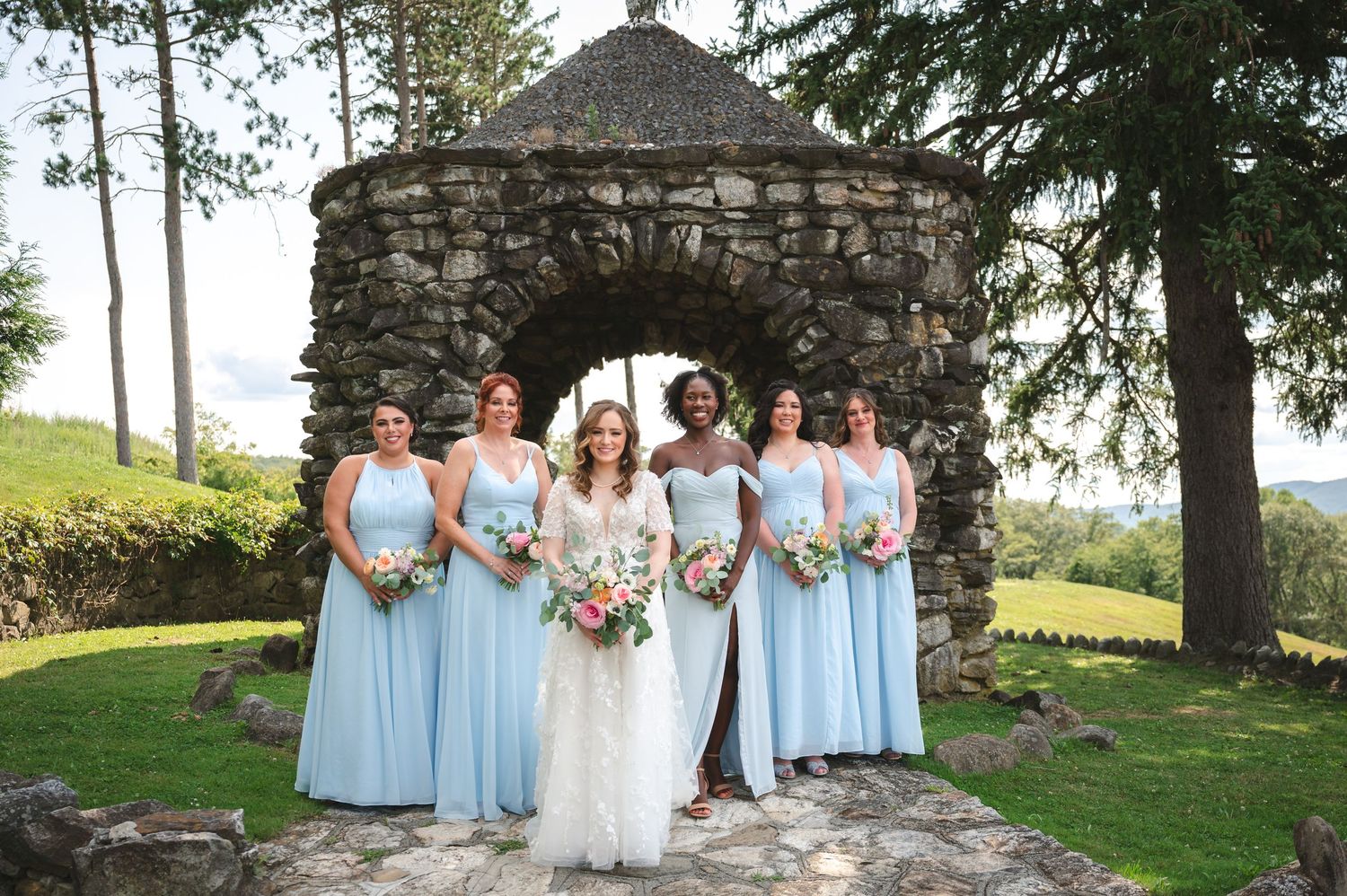Bride and wedding party in blue dresses pose smiling in front of stone gazebo on hilltop at The Garrison in Garrison, NY, Hudson Valley.