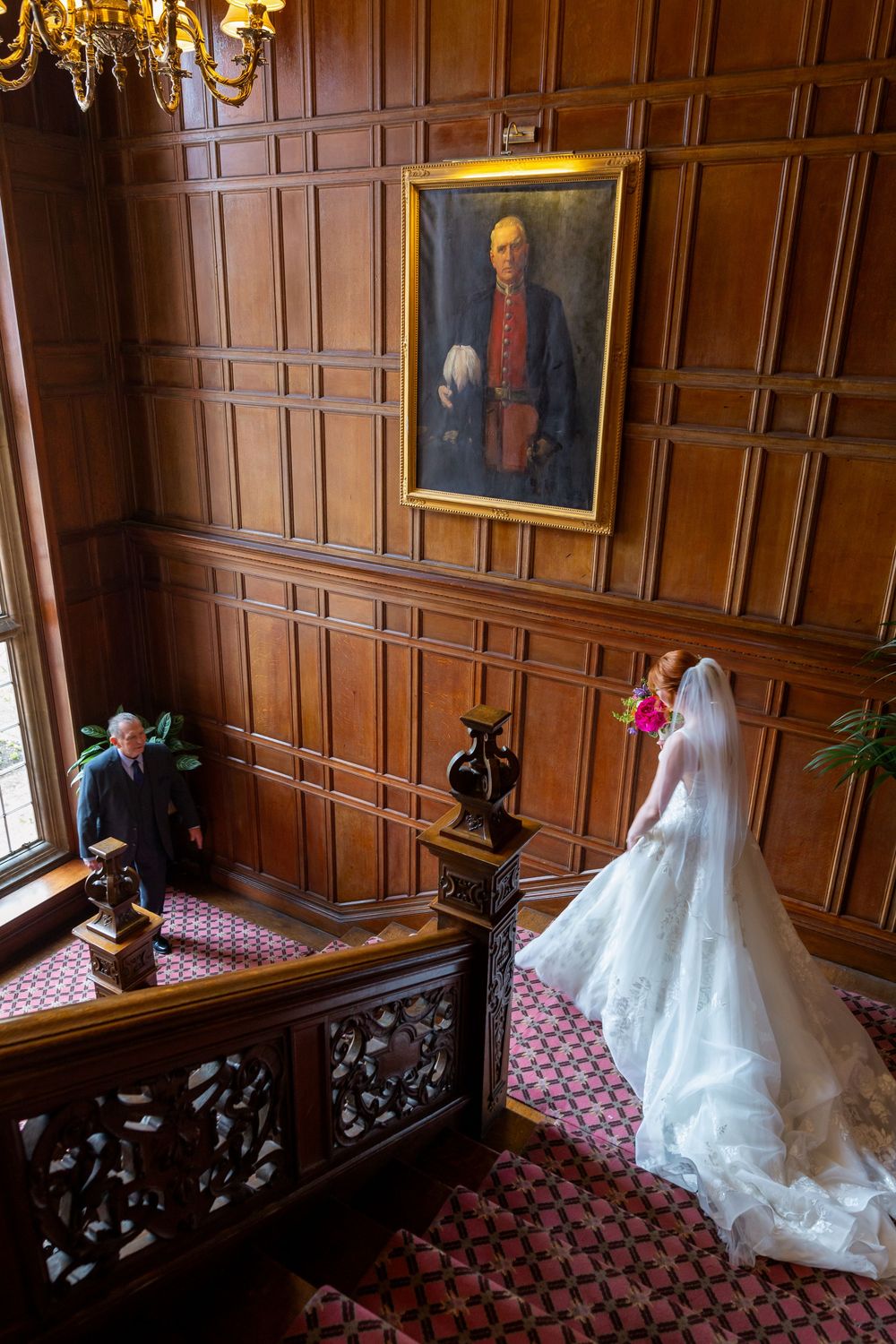 A bride in a flowing white gown stands on an elegant staircase with wood-paneled walls and a vintage portrait above.