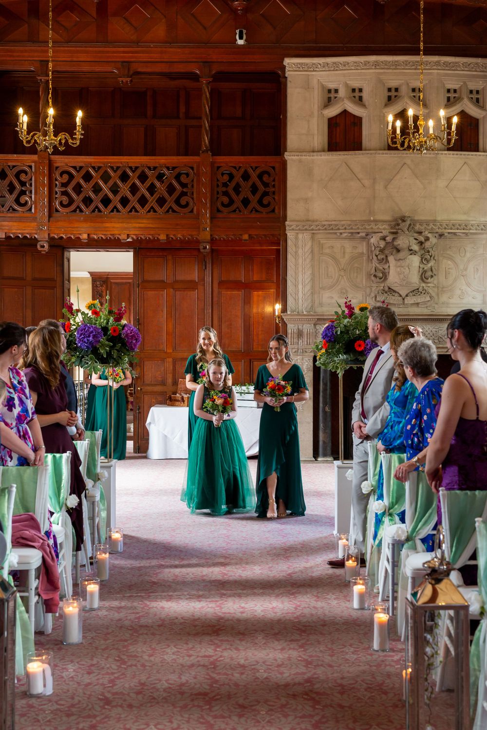 Wedding ceremony in an elegant wood-paneled hall with emerald green bridesmaids dresses and candlelit aisle decor.