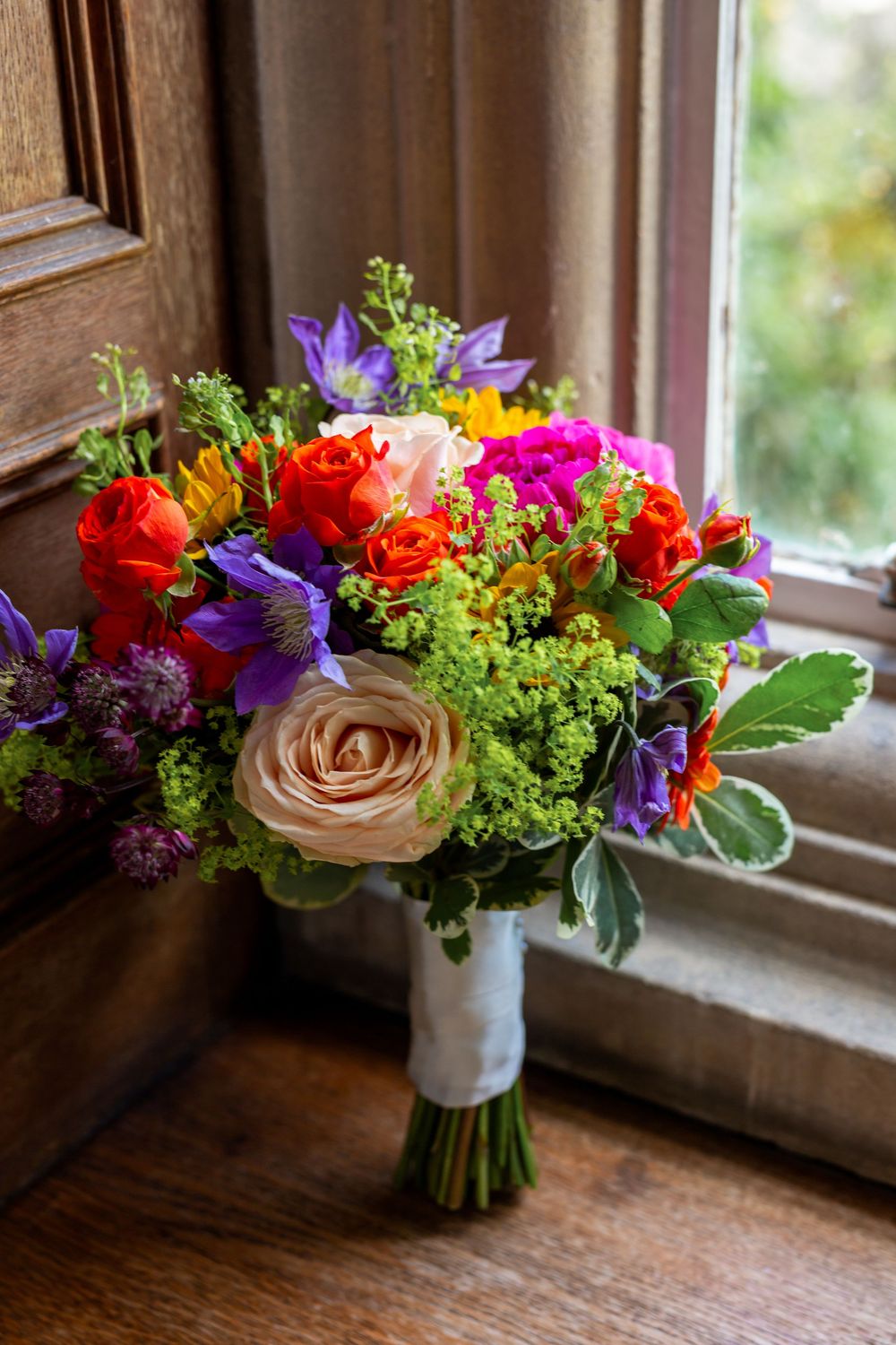 Vibrant wedding bouquet with orange roses, purple flowers and green foliage arranged on rustic wooden windowsill.