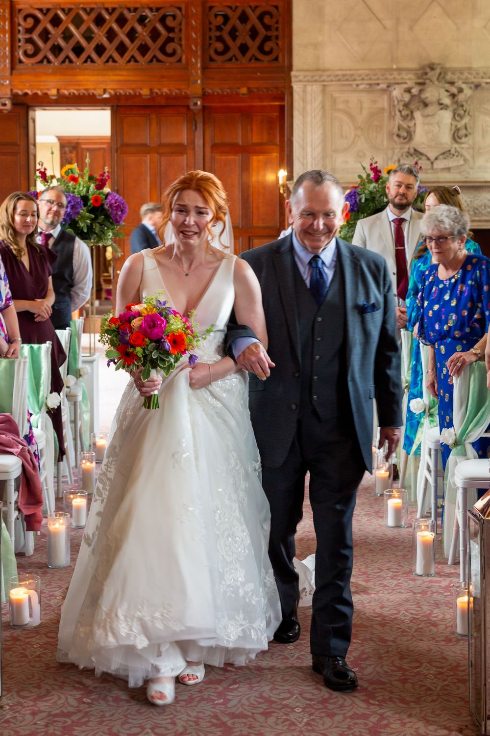 Wedding party walks down aisle in elegant venue decorated with colorful floral arrangements and candle-lit path.