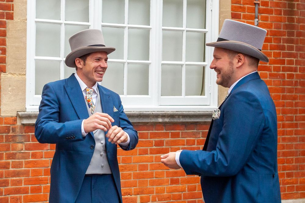 Two people in top hats and suits share a laugh while standing against a brick building exterior.